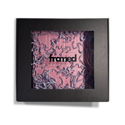 iepakojums, kas attēlo skaisti iepakotu magnētisko rāmi randiņu atmiņām. Attēlots mazais rāmis pārklāts ar dekoratīvam violetām skaidām, iepakots melnā kastē ar stikliņu vidū un logo FRAMED DATE